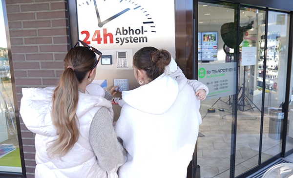 Zwei Frauen und ein Kind benutzen ein Tastenfeld an einem 24-Stunden-Abholsystem vor einer Apotheke. Eine Frau wartet mit dem Kind auf dem Arm, während die andere das Tastenfeld bedient, um den Lieferservice in der Nähe des Glaseingangs zu erreichen.