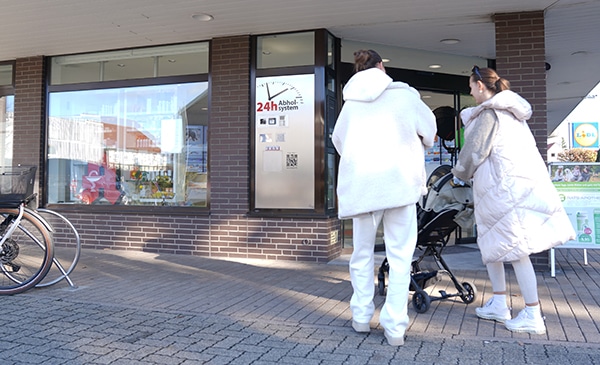 Zwei Personen in hellen Wintermänteln stehen vor einem Backsteingebäude mit großen Fenstern und unterhalten sich neben einem Kinderwagen. In der Nähe sind Fahrräder geparkt, und auf einem Schild mit einer Uhr steht "24h Abholsystem für Lieferservice".