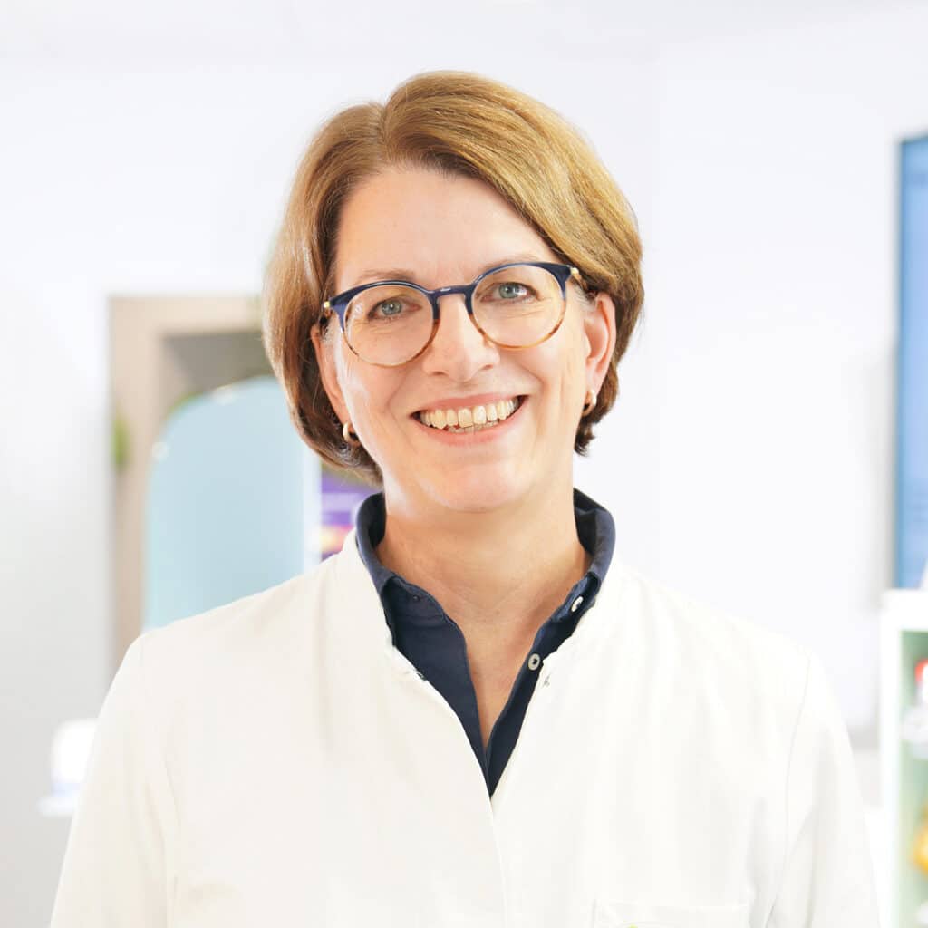 Eine Frau mit kurzen hellbraunen Haaren und Brille, die einen weißen Laborkittel und ein marineblaues Hemd trägt, lächelt in einer hell beleuchteten, modernen medizinischen oder klinischen Umgebung in die Kamera.