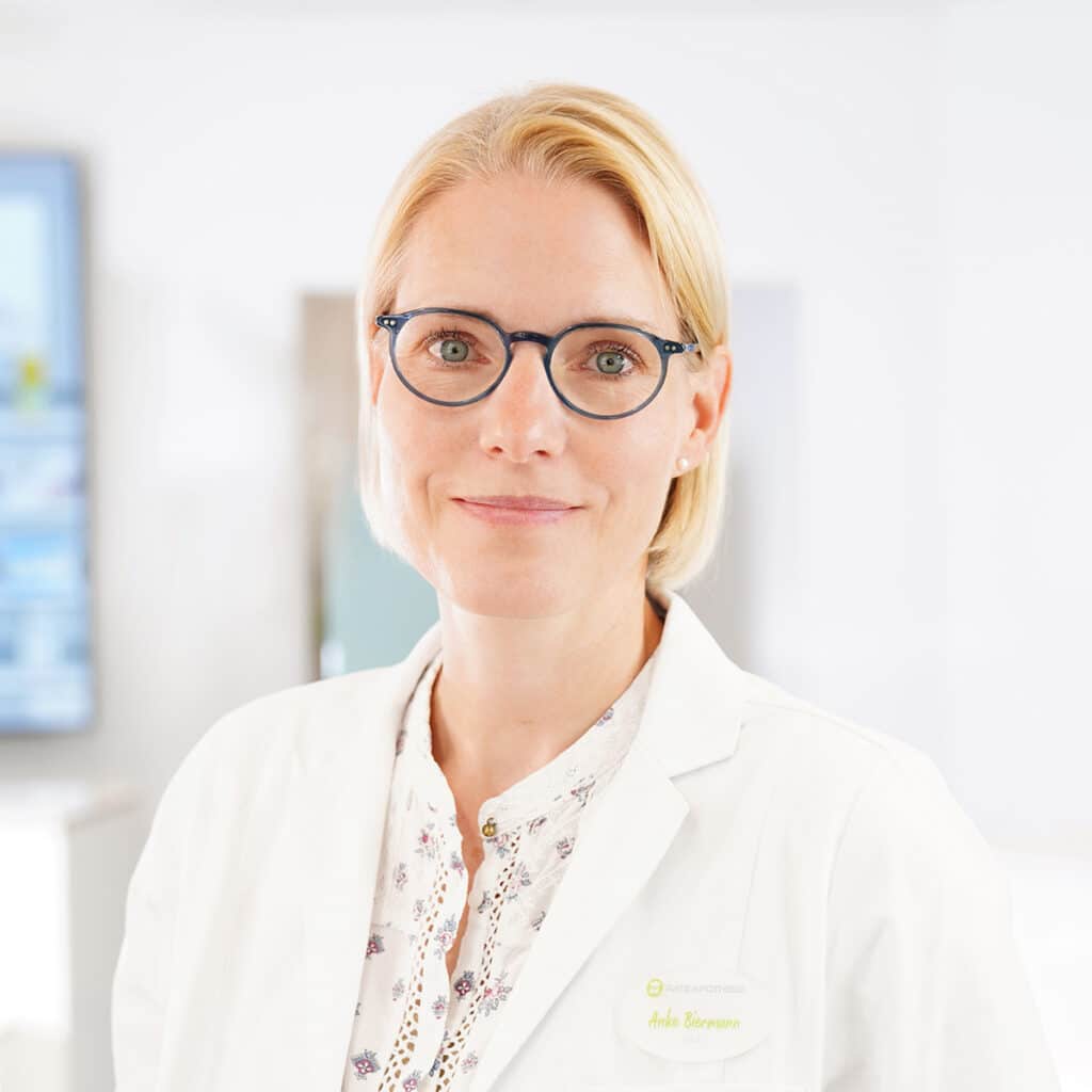 Eine Frau mit kurzen blonden Haaren und Brille, die einen weißen Laborkittel und eine gemusterte Bluse trägt, steht in einer hellen, modernen Arztpraxis. Sie blickt in die Kamera und lächelt sanft.