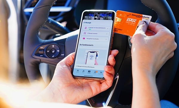 Eine Person sitzt in einem Auto, hält in der einen Hand ein Smartphone mit einer geöffneten Gesundheits-App und in der anderen eine deutsche Gesundheitskarte, während das Lenkrad sichtbar ist - bereit, Medikamente über den Lieferservice zu bestellen.