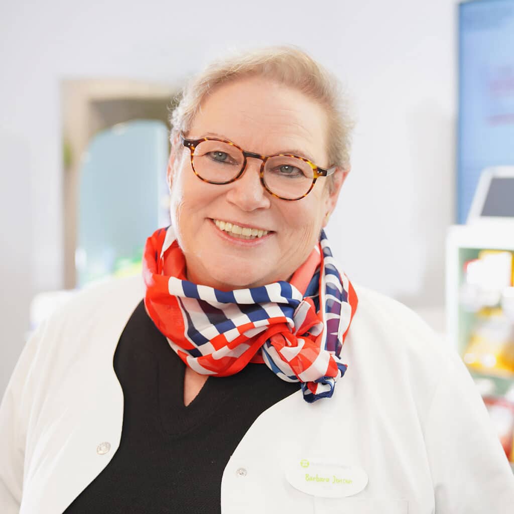 Eine lächelnde Frau mit Brille, weißem Mantel und einem rot-weiß-blau karierten Schal steht in einem Haus. Sie hat kurze Haare und trägt ein Namensschild mit der Aufschrift "Barbara Dantor".