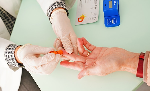 Eine medizinische Fachkraft mit Handschuhen sticht mit einem kleinen Gerät in den Finger eines Patienten für eine Blutuntersuchung; auf dem Tisch liegt medizinisches Material.