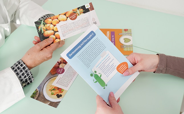 Zwei Menschen sitzen an einem Tisch und betrachten bunte Rezeptkarten mit Fotos von Lebensmitteln und Texten - ein einladender Moment aus einer Darmberatung. Nur ihre Hände sind zu sehen; der eine trägt einen weißen Ärmel, der andere ein kariertes Hemd.