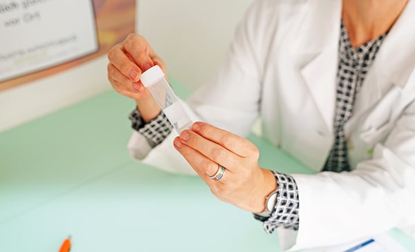 Eine Person in einem weißen Laborkittel hält ein durchsichtiges Plastikgefäß mit weißem Deckel in der Hand und sitzt an einem hellen Schreibtisch mit Unterlagen und einem Stift - eine ideale Umgebung für die Darmberatung.