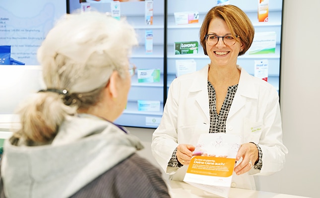 Ein lächelnder Apotheker im weißen Kittel hält einen Beipackzettel in der Hand, während er einer älteren Frau mit grauem Haar an einem Apothekentresen hilft. Im Hintergrund sind Regale mit Medikamentenschachteln zu sehen.