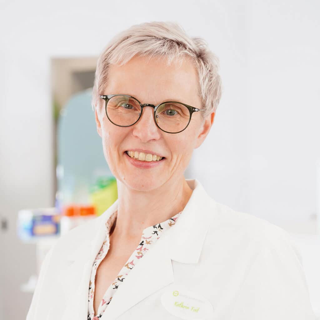 Eine lächelnde Frau mit kurzen grauen Haaren und Brille trägt einen weißen Laborkittel und ein Namensschild. Sie steht in einer hellen, sauberen Umgebung mit verschwommenen Regalen und Gegenständen im Hintergrund.