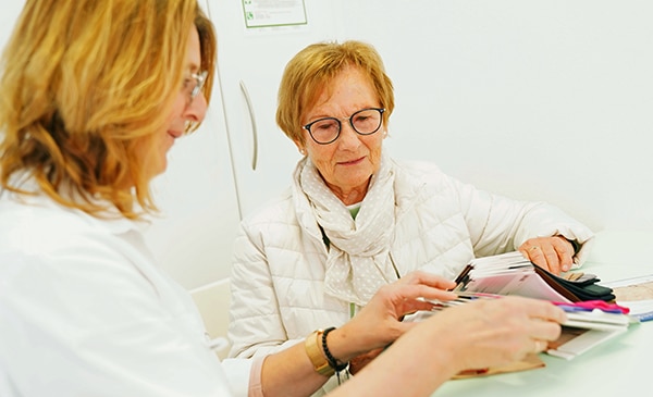 Zwei Frauen sitzen an einem Tisch und sehen sich farbige Stoffmuster für Kompressionsstrümpfe an. Die eine, die eine Brille und eine weiße Jacke trägt, beobachtet, während die andere die Muster erklärt. Beide scheinen in ein Gespräch vertieft zu sein.