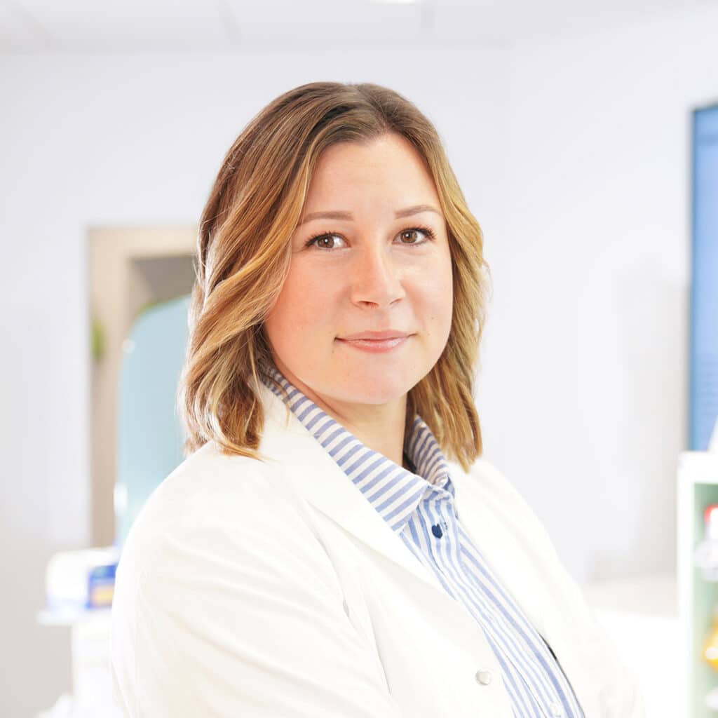 Eine Frau mit schulterlangem hellbraunem Haar, weißem Laborkittel und blau gestreiftem Hemd steht in einem hellen, modernen Büro und schaut selbstbewusst in die Kamera.