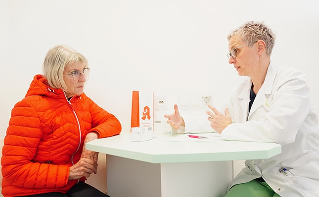 Eine ältere Frau in einer orangefarbenen Jacke sitzt an einem Tisch und hört einer medizinischen Fachkraft während einer Medikationsberatung aufmerksam zu, während medizinische Gegenstände auf dem Tisch ausgebreitet werden.
