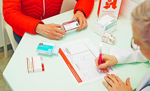 Eine Person in einer roten Jacke hält während einer Medikationsberatung Medikamentenschachteln, während eine medizinische Fachkraft an einem Tisch mit weiteren Medikamentenpackungen ein Formular ausfüllt.