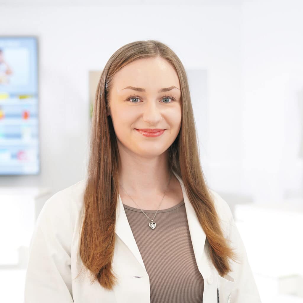 Eine junge Frau mit langen glatten braunen Haaren trägt einen weißen Laborkittel und eine Halskette mit einem Herzanhänger. Sie steht in einer hellen, modernen und verschwommenen professionellen Umgebung und lächelt in die Kamera.