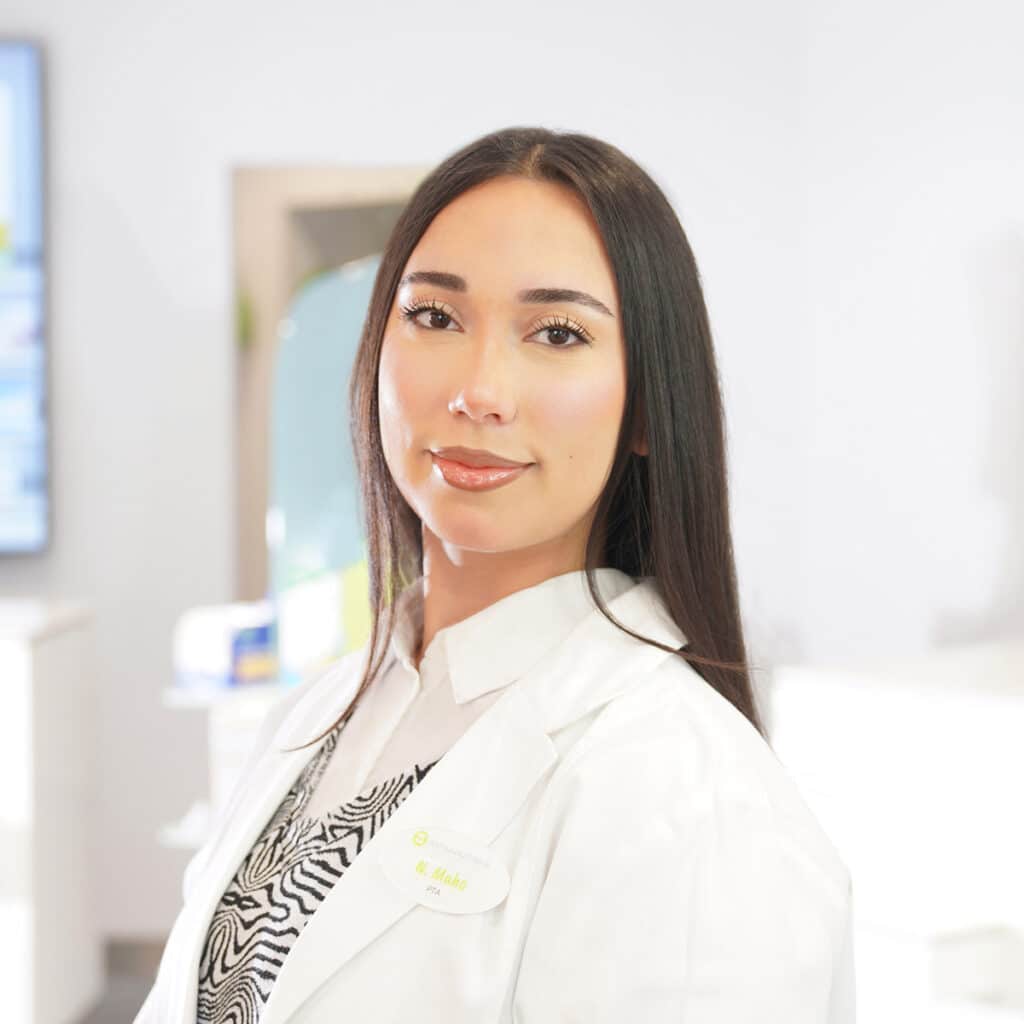 Eine Frau mit langen dunklen Haaren, die einen weißen Laborkittel und ein bedrucktes Oberteil trägt, steht in einer hellen, modernen medizinischen oder Laborumgebung und blickt selbstbewusst in die Kamera.