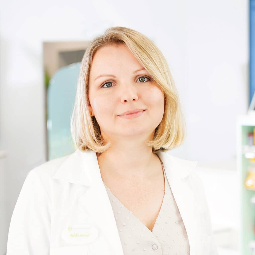 Eine Frau mit blondem Haar, die einen weißen Laborkittel und eine helle gemusterte Bluse trägt, steht in einem Haus und lächelt sanft in die Kamera.