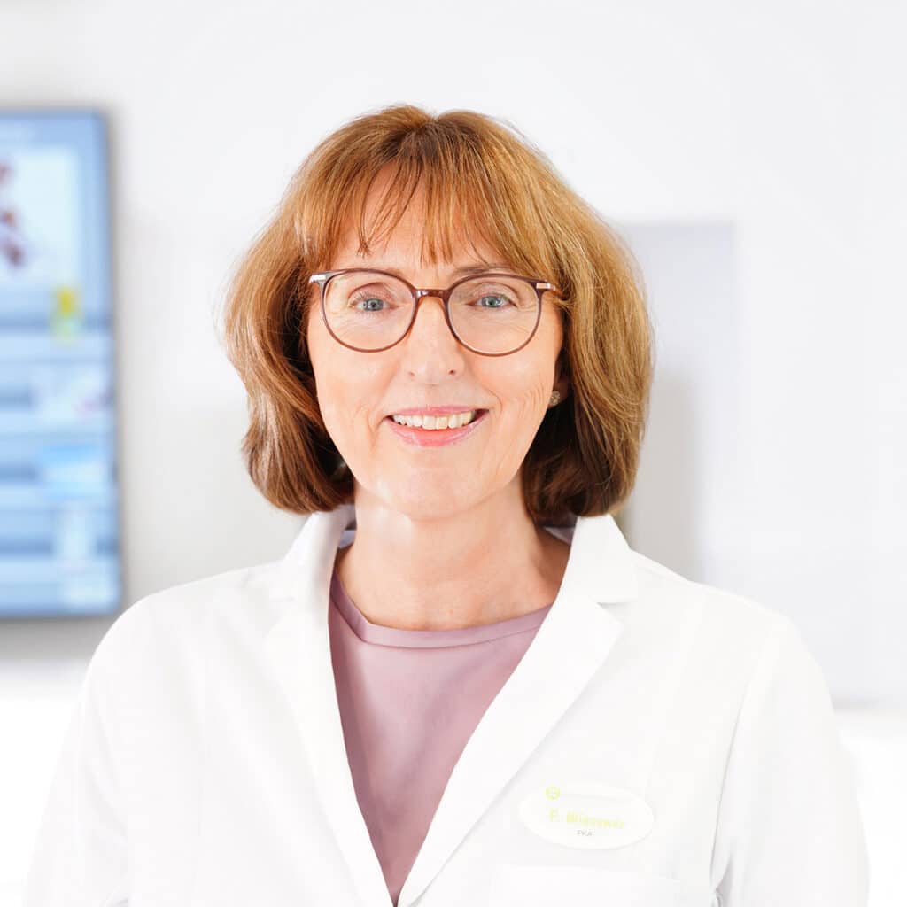 Eine Frau mit kurzen hellbraunen Haaren, Brille und weißem Laborkittel lächelt in die Kamera. Sie steht in einer hellen, modernen, klinischen Umgebung mit einem unscharfen Bildschirm im Hintergrund.