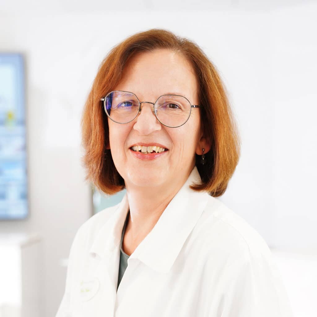 Eine Frau mit kurzen braunen Haaren und Brille lächelt, während sie einen weißen Laborkittel trägt und in einem hellen, modernen Büro oder einer medizinischen Umgebung steht.