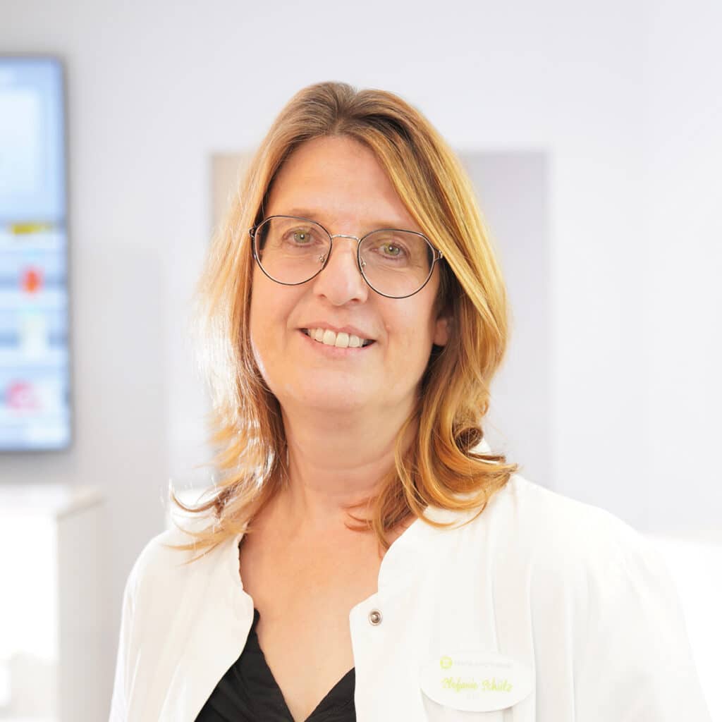 Eine Frau mit hellbraunem Haar und Brille, die einen weißen Kittel und ein Namensschild trägt, steht lächelnd in einer hellen, modernen Inneneinrichtung.