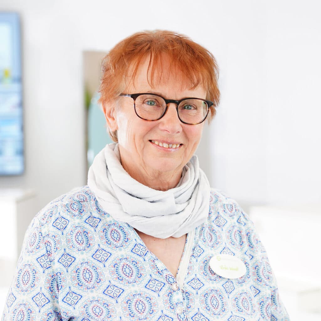 Lächelnde ältere Frau mit kurzen, hellroten Haaren und Brille, die ein gemustertes Hemd, einen hellen Schal und ein Namensschild trägt, steht in einer hellen, modernen Inneneinrichtung.