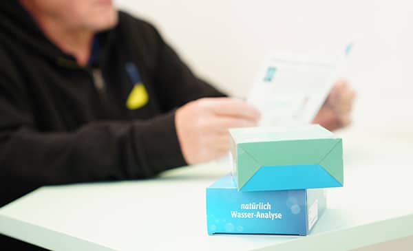 Eine unscharfe Person sitzt an einem Tisch und liest eine Zeitung. Im Vordergrund sind zwei Kartons gestapelt, der sichtbare ist mit "Natürlich Wasser-Analyse" beschriftet.